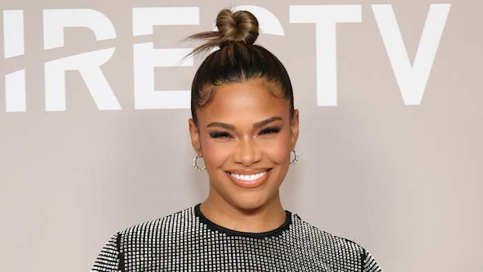 Kamie Crawford poses in a bedazzled black dress and a top knot and smiles for the camera.